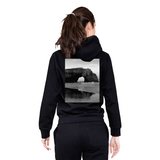 Person wearing a black hoodie with a scenic print on the back, standing against a white background